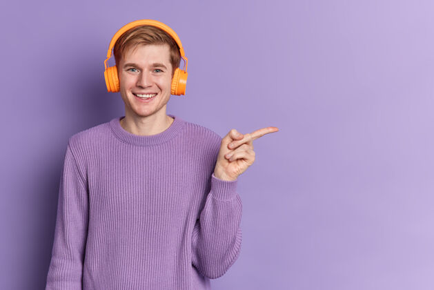 娱乐积极的男性青少年 蓝眼睛 快乐的微笑 穿着休闲的紫色毛衣 戴着立体声耳机听音乐 在拷贝空间上指指点点 在拷贝空间上做广告年轻人的爱好科技男人收音机