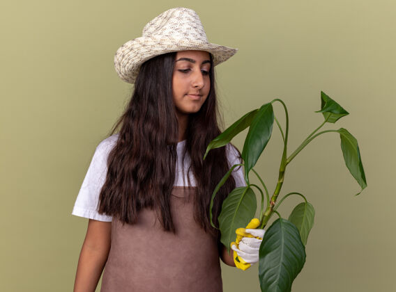 花园穿着围裙 戴着夏帽 戴着工作手套的年轻园丁女孩 手拿植物 面带微笑地看着它站在绿色的墙上穿上微笑帽子