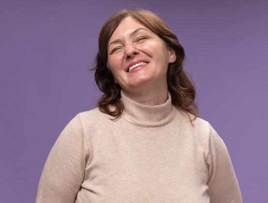 中年身穿米色套头衫的中年妇女站在紫色的墙上 幸福快乐地闭着眼睛微笑着高领毛衣站着年龄