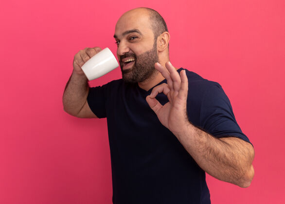杯子一个留着大胡子的男人穿着海军蓝t恤 手里拿着一杯茶 正准备喝一杯 在粉红色的墙上 他站着一个好的标志胡子站起来好的