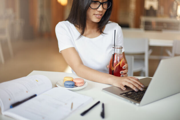 眼镜女人很聪明年轻的女人在咖啡馆里一边用笔记本电脑喝柠檬水 一边等着吃麦卡龙糕点休闲笔记本电脑大学