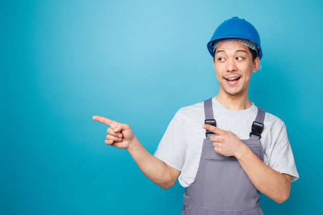蓝色兴奋的年轻建筑工人戴着安全帽和制服看着并指着旁边安全侧线穿