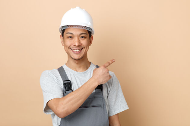 建筑微笑着的年轻建筑工人戴着安全帽和制服指着角落穿指向空间
