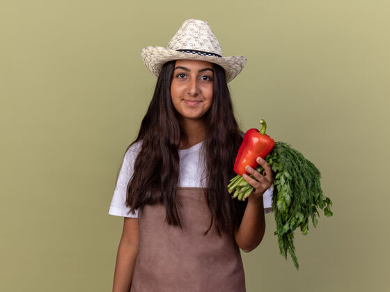 帽子戴着围裙和夏帽的快乐的年轻园丁女孩手持新鲜的红甜椒和新鲜的草药微笑着站在绿色的墙上胡椒香草年轻