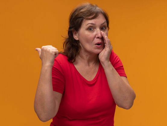 拇指穿着红色t恤的中年妇女低声说着一个秘密 手放在嘴边 拇指指向后面 站在橙色的墙上指点年龄近