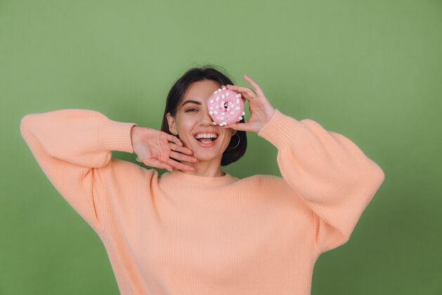 女孩穿着休闲桃色毛衣的年轻时尚女性 隔离在绿色橄榄色墙壁上 粉色甜甜圈快乐复制空间甜甜圈黑发肖像