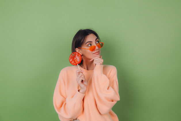 甜点年轻时尚的女士穿着休闲的桃色毛衣 戴着橘色眼镜 隔离在绿色橄榄色的墙上 用橘色棒棒糖若有所思地看着一边思考着复制空间五颜六色享受青少年