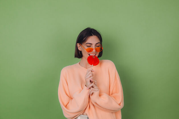 乐趣年轻时尚的女士穿着休闲的桃色毛衣 戴着橙色的眼镜 隔离在绿色橄榄色的墙上 带着橙色棒棒糖的正面微笑复制空间表情享受漂亮