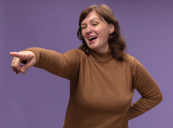 指向快乐快乐的中年妇女穿着棕色高领毛衣 一边看着一边用食指指着紫色墙壁上的东西手指东西中间