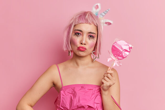 表情心烦意乱的粉色头发的亚洲年轻女子撅起嘴唇已经漏了妆看着镜头被冒犯有人拿着裹着的糖果穿着时髦的裙子不满意鲍勃女性