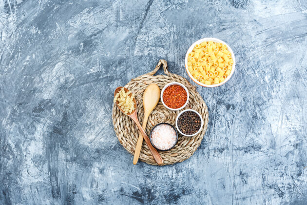 酱汁碗里的什锦意大利面和带香料的木勺灰色石膏和柳条垫子背景上的俯视图传统自制午餐