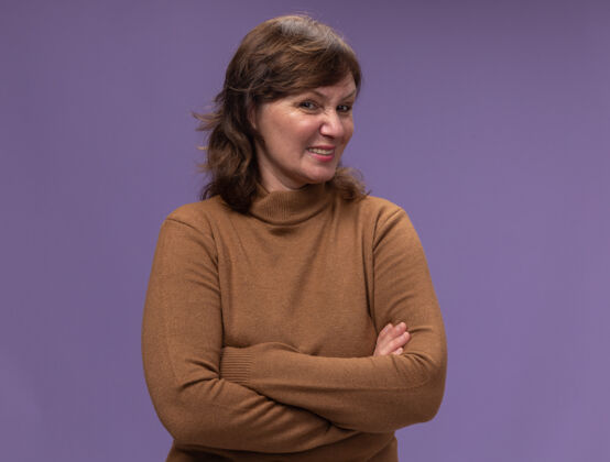 女人身穿棕色高领套头衫 双臂交叉 表情怀疑的中年妇女站在紫色的墙上中年站姿高领毛衣