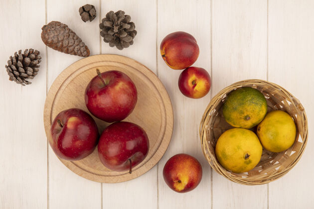 特写顶视图红色的苹果放在木制的厨房板上 橘子放在桶上 桃子和松果被隔离在白色的木制表面上桶营养锥