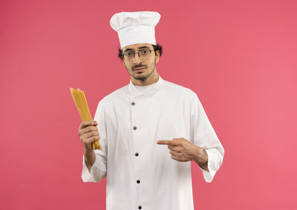 男年轻的男厨师穿着厨师制服 戴着眼镜拿着并指着意大利面点拿着烹饪