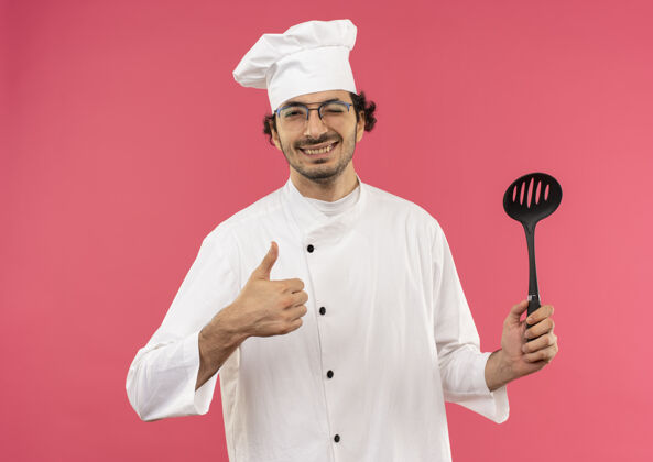 男面带微笑的年轻男厨师穿着厨师制服 戴着眼镜 举着抹刀 拇指朝上眼镜穿向上