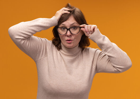 高领毛衣穿着米色高领套头衫 戴着眼镜的中年妇女站在橘色的墙上惊讶不已站年龄中年