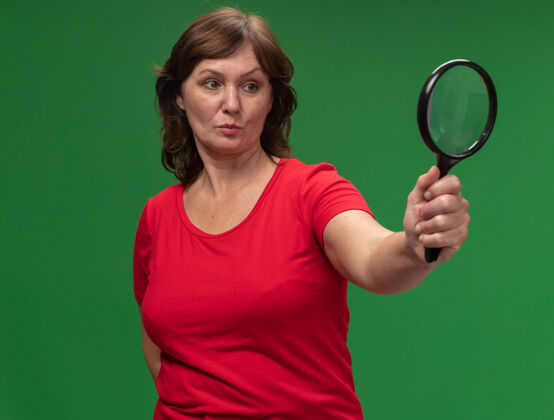中年身穿红色t恤的中年妇女举着放大镜 严肃地站在绿色的墙上看着它放大镜年龄镜子