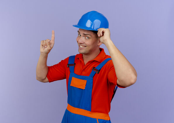 建设者年轻的男建筑工人穿着制服 戴着安全帽 手放在头盔上 指着上面年轻安全印象