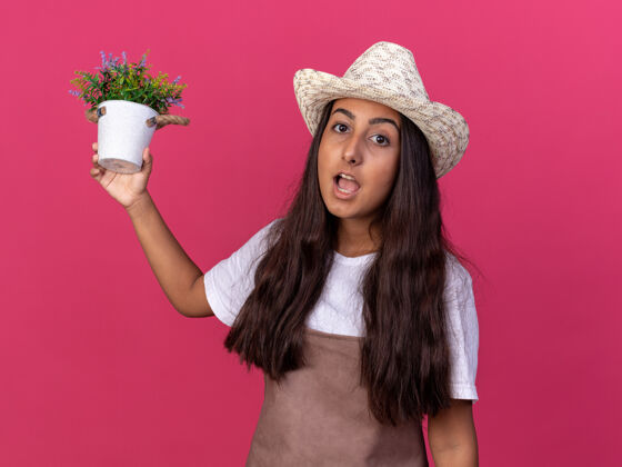 帽子穿着围裙 戴着夏帽 手里拿着盆栽植物的年轻园丁女孩站在粉红色的墙上惊讶不已锅立场年轻的