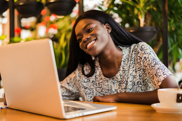 年轻坐在咖啡馆里用笔记本电脑工作的年轻非洲女人杯子打字自由职业者