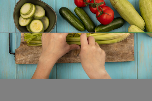 餐女性用刀在木制厨房板上切芹菜的手的俯视图 西红柿 黄瓜和西葫芦被隔离在蓝色的木制表面上素食厨房女性