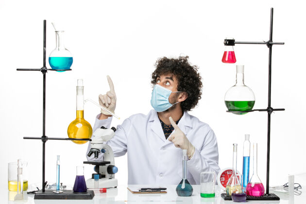 浅白色正面图：男药剂师穿着医疗服 戴着口罩 坐在不同的溶液中 在白色的空间里摆姿势实验室只是西装