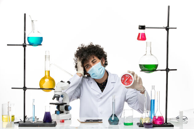 实验室外套正面图：男药剂师穿着医疗服 戴着面罩 白色桌子上拿着红色的钟科学面具实验室