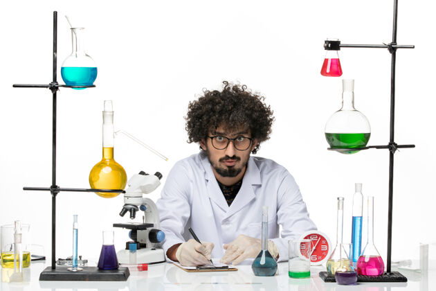 书桌正面图：身着医疗服的男性化学家在白色办公桌上写笔记写作正面实验室