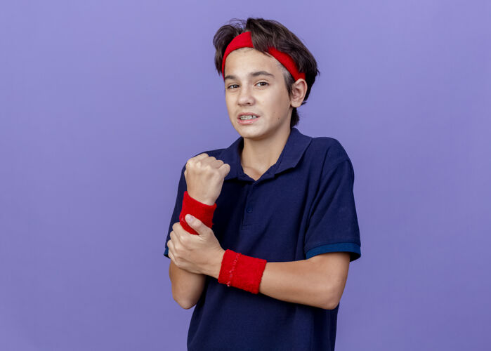 年轻疼痛的年轻帅气的运动男孩戴着头带和带牙套的腕带 把手腕隔离在紫色的墙上 留着复制空间手腕牙科男孩
