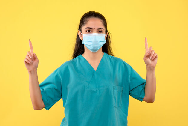 保护盖美丽的女医生 戴着医用面罩 手指指向黄色的墙上医生盖外套