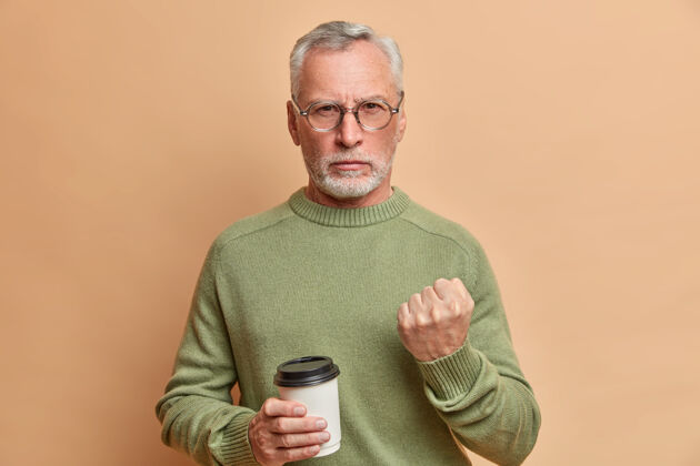 人严厉愤怒的大胡子老人严肃地看着前面试图警告你拿着一次性咖啡杯穿着休闲套头衫对着棕色墙壁摆姿势拳头疯狂欧洲人