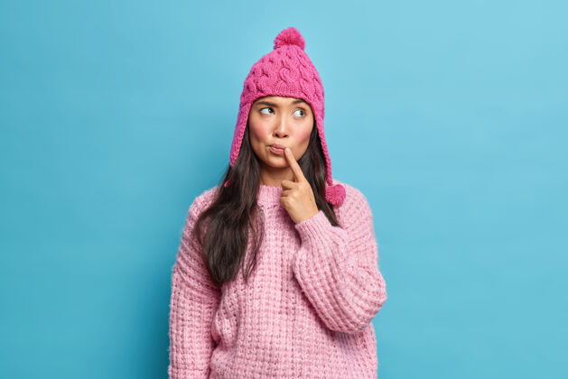 羊毛美丽自然的亚洲年轻女子 穿着针织毛衣 戴着帽子 手指紧靠嘴唇 专注地放在一边 想着被隔离在蓝色墙壁上的寒假计划思维室内肖像