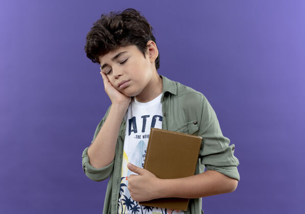小学生闭上眼睛 生病的小男孩拿着书 把手放在隔离在紫色墙上的脸颊上病了脸颊小