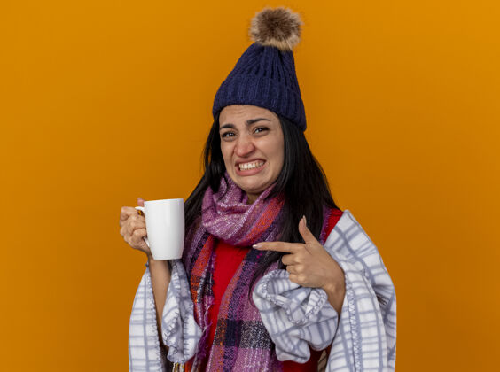 包装恼怒的年轻白种人生病的女孩戴着冬天的帽子 围着格子呢围巾 拿着茶杯指着橘黄色的墙壁上的复制空间年轻茶穿