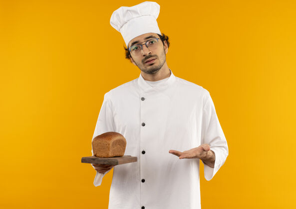 制服未出狱的年轻男厨师身穿厨师制服 手持眼镜 用手点着胡子 把砧板隔离在黄色的墙上穿年轻不愉快