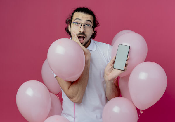 抱着惊讶的帅哥戴着眼镜站在气球中间拿着电话把手放在脸颊上隔离在粉红色的墙上男人脸颊中间