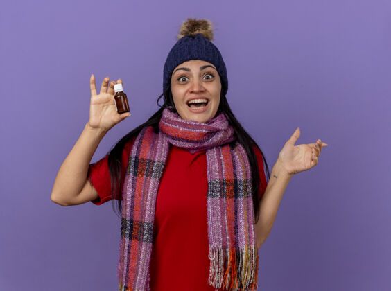 围巾快乐的年轻白种女孩戴着冬天的帽子和围巾 手里拿着药水 指着隔离在紫色墙上的一面壁板穿帽子