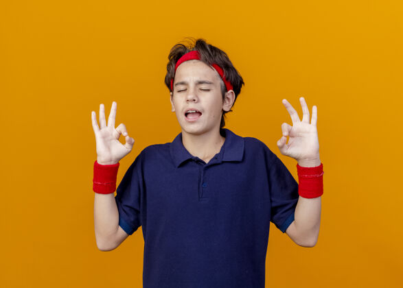 封闭自信的年轻帅气的运动男孩戴着头带和护腕 戴着牙套 闭着眼睛在橙色的墙上做着“ok”的手势橙色帅气年轻
