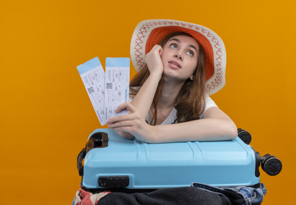 胳膊体贴的年轻旅行家女孩戴着帽子拿着飞机票 把手臂放在箱子上孤零零的橙色墙上女孩手提箱飞机