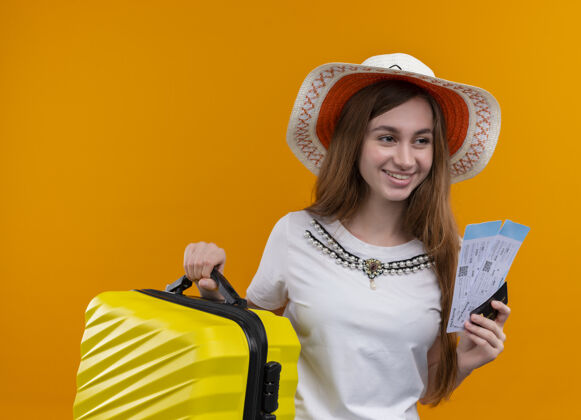 信用证带着微笑的年轻旅行家女孩戴着帽子拿着手提箱和机票 信用卡贴在孤立的橙色墙上女孩机票持有