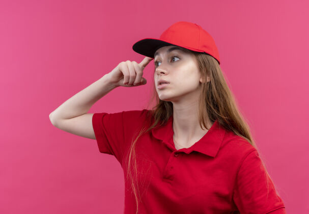 手指体贴的年轻送货员穿着红色制服 手指放在头上 看着孤立的粉红色墙上的左边年轻分娩制服