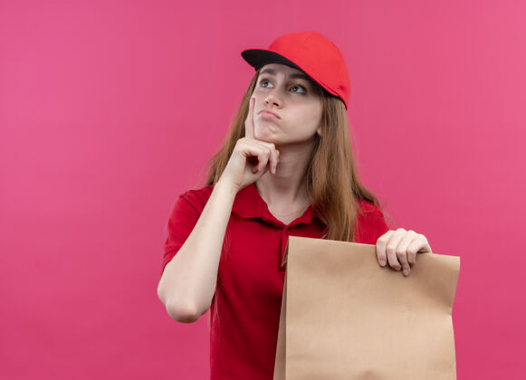 交货体贴的年轻送货女孩 穿着红色制服 手拿纸袋 把手放在下巴下面 放在隔离的粉色墙壁上 留着复印空间年轻女孩思想