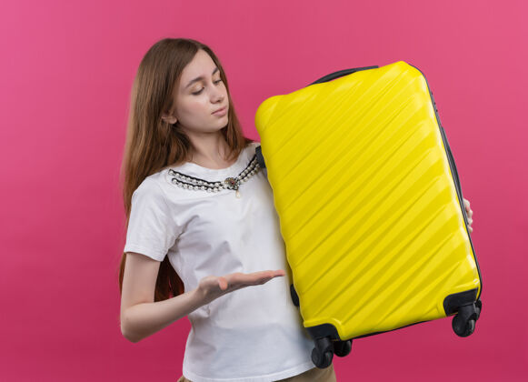 女人年轻的旅行家女孩拿着手提箱 用手指着孤零零的粉红色墙上的手提箱年轻人拿着手提箱