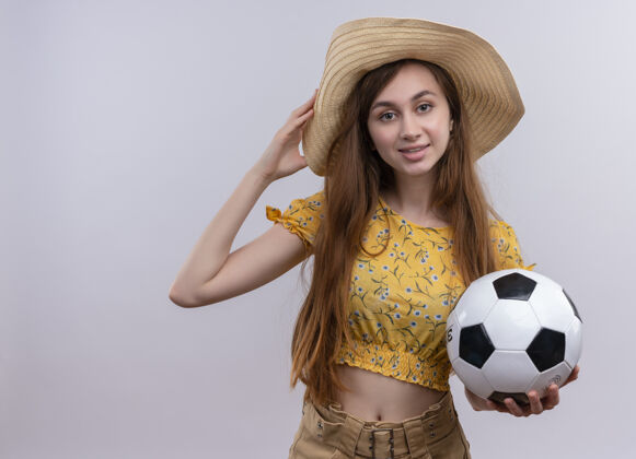 足球微笑的年轻女孩戴着帽子拿着足球把手放在帽子上孤立的白色墙壁与复制空间球抱着帽子