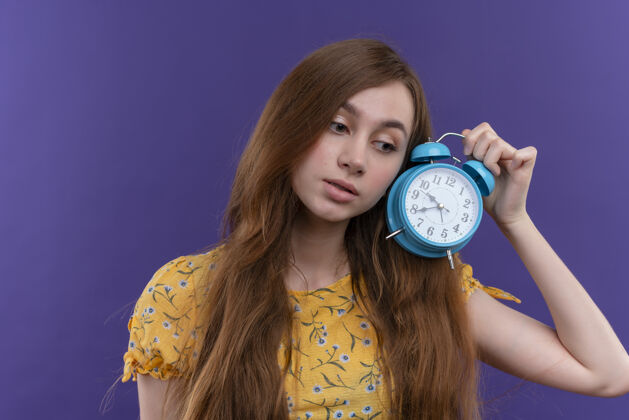 女孩一个体贴的年轻女孩 把闹钟放在孤零零的紫色墙上年轻闹钟抱着