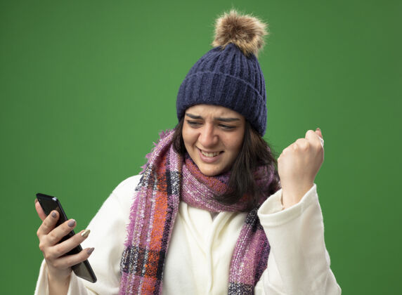长袍快乐的年轻白种人生病的女孩穿着长袍 戴着冬天的帽子 戴着围巾 手拿餐巾 拿着手机 看着手机做着“是”的手势 隔离在绿色的墙上穿电话女孩