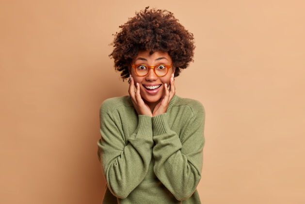 眼镜快乐的非洲头发女人双手放在脸颊上微笑着高兴地看着前面戴着眼镜和毛衣隔离在棕色的墙上女士套头衫夏天