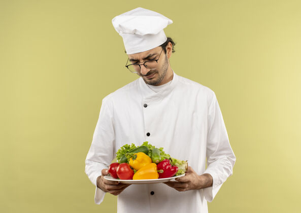 思考年轻的男厨师穿着厨师制服 戴着眼镜 拿着蔬菜 看着隔离在绿墙上的盘子佩戴厨师盘子