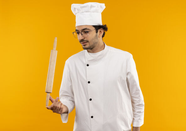 眼镜高兴的年轻男厨师穿着厨师制服 戴着眼镜 手指上拿着擀面杖 隔离在黄色的墙上滚烹饪厨师