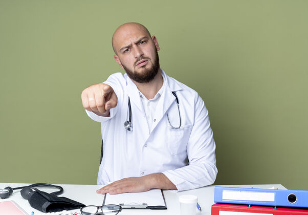 医生严格年轻的秃头男医生穿着医用长袍和听诊器坐在办公桌上用医疗工具显示你的手势隔离在绿色的墙上严格手势坐着